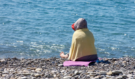 A woman sits on a beach with her feet in the water. She is wearing a yellow blanket and a red hat. The scene is peaceful and relaxing, with the sound of the waves and the warmth of the sun on her skinの写真素材