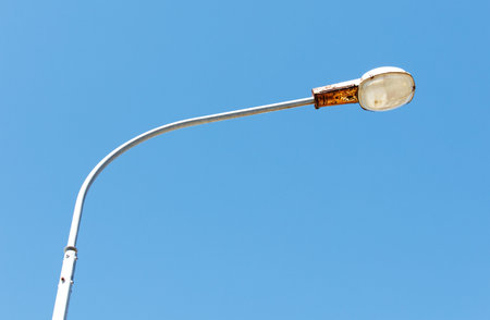 A street light is hanging from a pole. The light is rusted and the pole is bent. The sky is clear and blueの写真素材