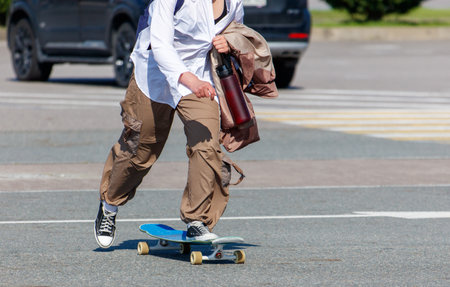 A man is riding a skateboard down the street. He is wearing a white shirt and brown pants. He is carrying a backpack and a water bottleの写真素材