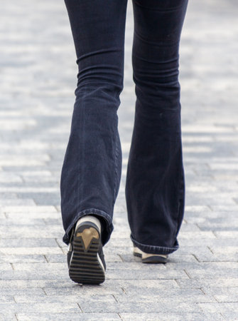 A woman wearing blue jeans and a black shoe walks down a brick sidewalk. Concept of casualness and leisure, as the woman is taking a leisurely strollの写真素材