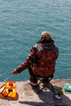 A man is fishing in the ocean. He is wearing a red jacket and sitting on a chairの写真素材