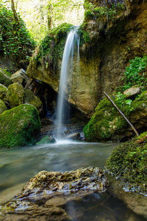A waterfall is flowing into a small pool of water. The water is clear and the rocks surrounding the pool are mossy. The scene is peaceful and sereneの写真素材