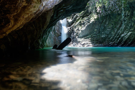 A large cave with a waterfall and a small stream. The water is clear and the sunlight is shining through the caveの写真素材