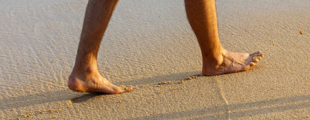 A man is walking on the beach barefoot. The sand is warm and the water is calm. The man's feet are visible in the sand, and he is enjoying the peaceful atmosphereの写真素材
