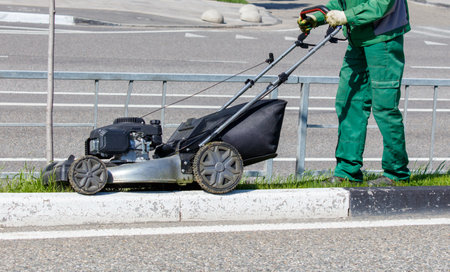 A man in a green outfit is pushing a lawn mower. The mower is on the grass next to a curbの写真素材