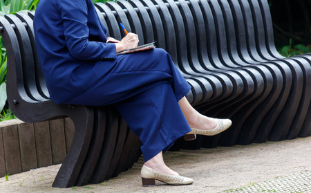 A woman sits on a bench with a pen and notebook. She is writing something down. The bench is made of metal and is longの写真素材