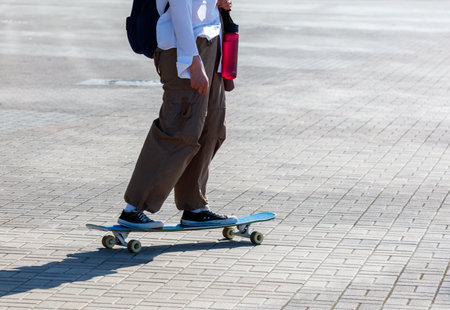 A person is walking on a sidewalk with a skateboard. The person is wearing a backpack and a red water bottleの写真素材