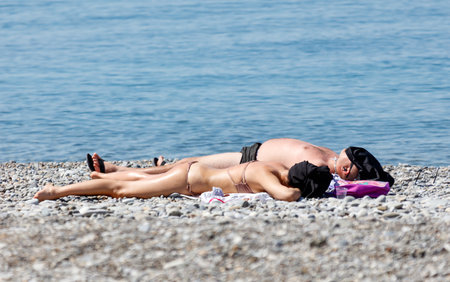 A man and a woman are laying on the beach, enjoying the sun and the water. The man is wearing a black shirt and the woman is wearing a bikini. Scene is relaxed and carefreeの写真素材
