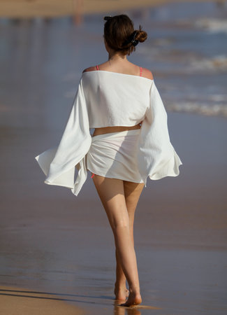A woman is walking on the beach wearing a white dress and a red top. She is barefoot and has her hair in a bunの写真素材