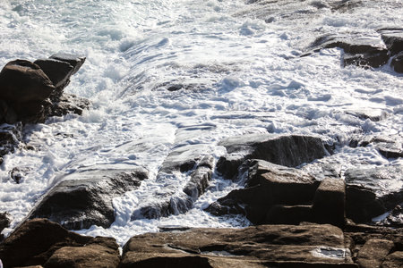 The ocean is rough and the waves are crashing against the rocks. The water is white and the rocks are grayの写真素材