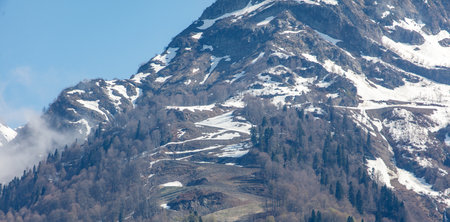 A mountain covered in snow with trees in the foreground. The snow is piled up on the mountain and the trees are bare. The sky is clear and the sun is shiningの写真素材