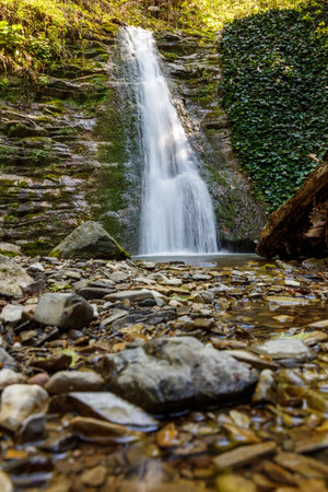 A waterfall is flowing down a rocky cliff. The water is clear and the rocks are scattered around the area. The scene is peaceful and serene, with the sound of the water creating a calming atmosphereの写真素材