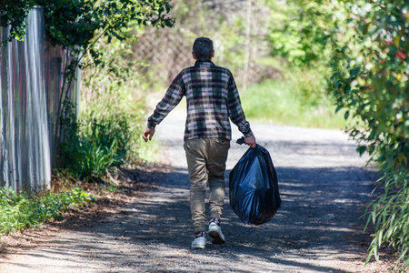 A man is walking down a road with a black trash bag in his hand. The scene is peaceful and quiet, with trees lining the path. The man seems to be in a hurryの写真素材
