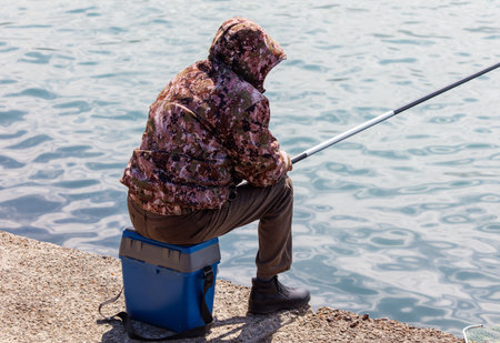 A man is sitting on a blue box while fishing. The man is wearing a jacket and pants