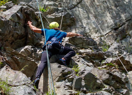 A man is climbing a rock wall with a yellow helmet on. He is wearing a blue shirt and black pantsの写真素材