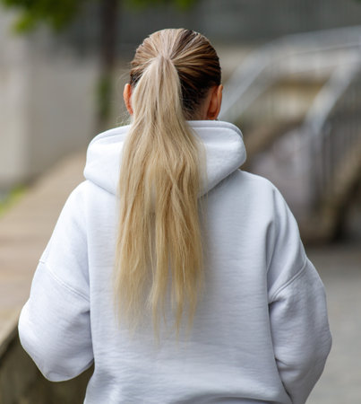 A blonde girl with long hair is wearing a white hoodie. She is standing on a sidewalkの写真素材