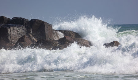 A large wave crashing against a rocky shore. The water is white and frothy, and the rocks are scattered throughout the scene. Scene is powerful and dramaticの写真素材