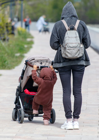 A woman is walking with a child in a stroller. The child is wearing a brown coat and is holding onto the stroller's handle. The woman is wearing a backpack and is looking at her phoneの写真素材