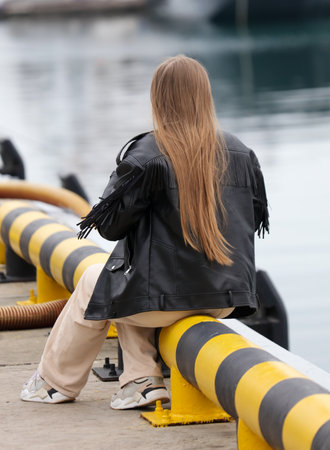 A woman with long hair is sitting on a yellow and black metal pole. She is wearing a black jacket with fringeの写真素材