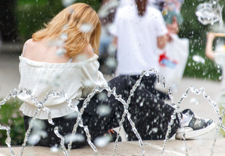 A woman is sitting on a bench next to a fountain. The water is spraying out of the fountain and the woman is looking at itの写真素材