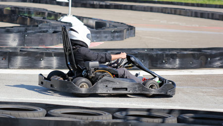 A man is driving a go kart on a track. The man is wearing a helmet and a black jacket. The go kart...