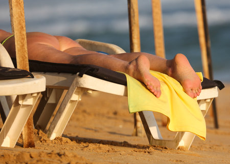 A person is laying on a beach chair with their feet up on a yellow towel. The beach chair is next to a pole and another chairの写真素材