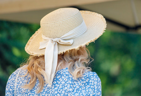 A woman wearing a straw hat with a white ribbon tied around it. The woman is wearing a blue dress and is standing in a green fieldの写真素材