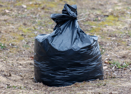 A black plastic bag is sitting on the ground. The bag is full and has a plastic bag tied to it. The bag is on a grassy areaの写真素材