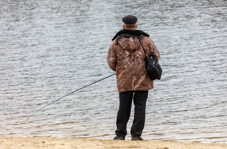 A man is fishing in a lake wearing a brown jacket and black pants. He is holding a fishing rod and a bagの写真素材