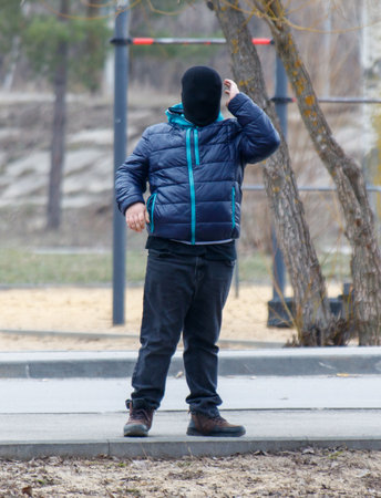 A man in a black hoodie and mask stands on a sidewalk. He is wearing blue and black jeans and has a black hat onの写真素材