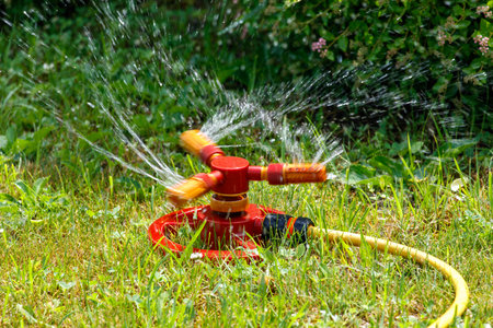 A red sprinkler with three nozzles is sitting on the grass. The water is spraying out of the sprinkler, creating a wet and refreshing atmosphereの写真素材