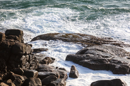 A rocky shoreline with a large wave crashing against the rocks. The water is white and frothy, and the rocks are scattered throughout the scene. Scene is one of power and strengthの写真素材