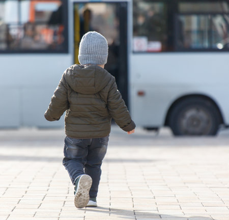 A young child wearing a gray hat and a green jacket is running down a sidewalk. The scene is set in a city, with a bus parked in the background. The child appears to be enjoying the outdoorsの写真素材