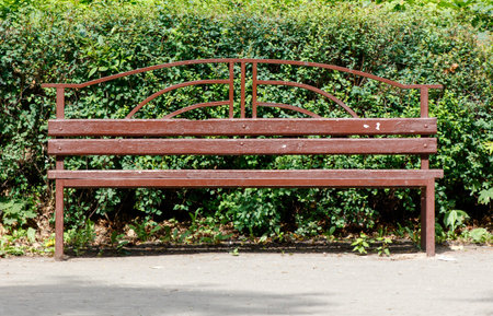 A brown bench is sitting in front of a bush. The bench is empty and has a weathered look. The bush is green and has a lot of leaves. The scene is peaceful and quietの写真素材