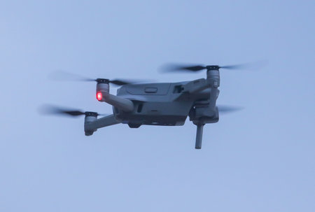 A small drone is flying in the sky. The drone is white and has red lights on the bottom. The sky is...