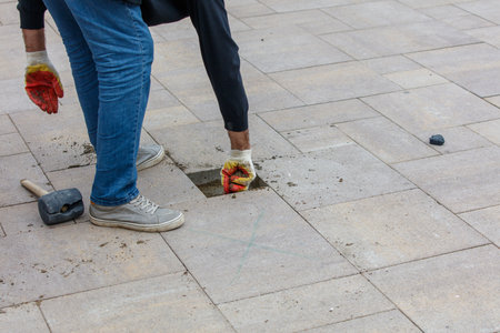 A man is working on a sidewalk, using a hammer and a chisel to fix a hole in the pavementの写真素材