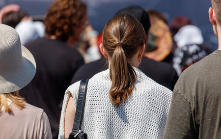 A woman with a ponytail and a white shirt is standing in a crowd of people. She is wearing a hat and a black purseの写真素材