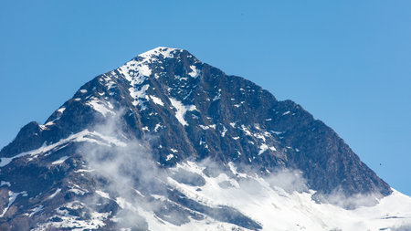 A mountain covered in snow with a blue sky in the background. The mountain is very tall and the snow...