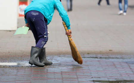 A young person is sweeping the sidewalk with a broom. The scene is set in a city, with several people walking around in the backgroundの写真素材
