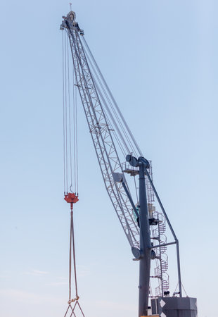 A crane is lifting a large object. The crane is tall and has a red and white crane hook. The sky is...