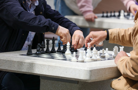 Two people playing chess on a table. One of the players is wearing a blue jacket. The other player...