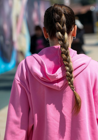 A girl with long hair and a pink hoodie. She has a ponytail and her hair is braided