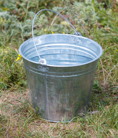 A silver bucket with a handle is filled with water. The bucket is sitting on the grass. The water in the bucket is clear and calmの写真素材
