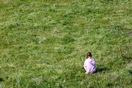 A girl is sitting in a field of grass. She is wearing a pink hoodie. The grass is green and the sky is blueの写真素材