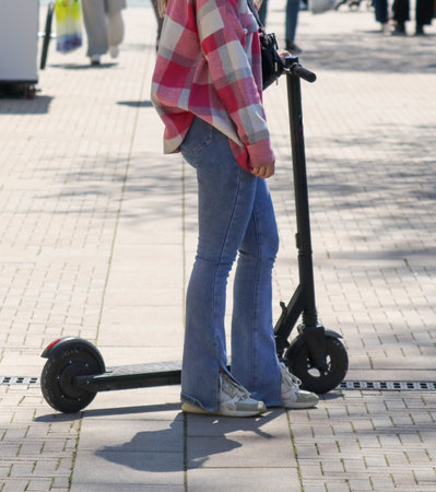 A woman is standing next to a black scooter. She is wearing a pink and white plaid jacket and blue jeansの写真素材