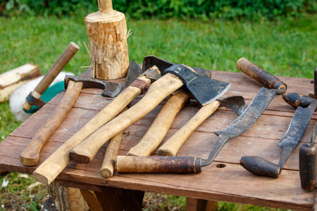 Table with a bunch of tools on it. There are five axes and three hammers. The axes are all different...
