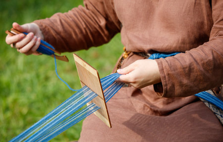 Person is sitting on the grass and is weaving a blue and white string. The person is wearing a brown...