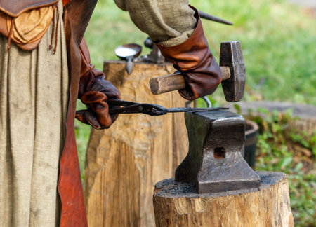 Man is hammering a piece of metal. He is wearing leather gloves. The metal is on a wooden block