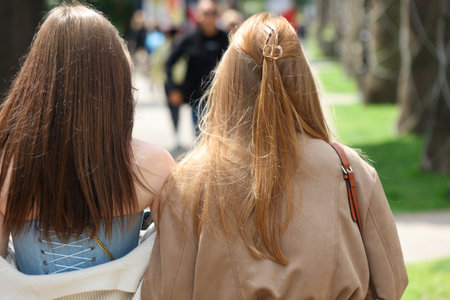 Two women with long hair are walking down a street. One of them has a brown jacket on