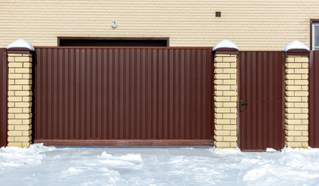 A brown fence with a gate in front of a brick building. The gate is closed. The fence is made of...